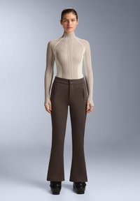 Haut beige côtelé à manches longues avec col montant, associé à un pantalon marron évasé et ajusté, doté de fermetures éclair à l'avant, d'une couture minimale et d'une fermeture à deux boutons.