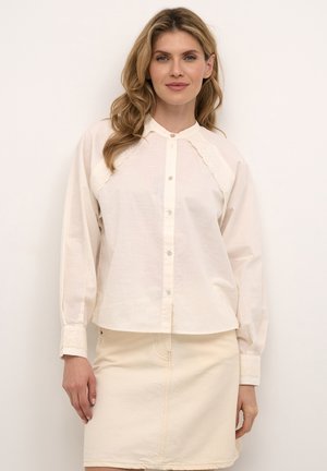 Lys beige skjorte med knapper og en blød tekstur, der har en rund krave og broderede blonde detaljer på skuldrene og manchetterne.