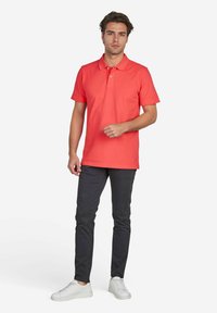 Rotes Polohemd mit kurzen Ärmeln und einer Knopfleiste mit zwei Knöpfen, kombiniert mit dunkelgrauen Slim-Fit-Hosen und weißen Sneakers.