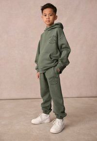 Zielona bawełniana bluza z kapturem, z kieszenią z przodu i nadrukowanym logo; dopasowane zielone spodnie dresowe z ściągaczami; w komplecie białe buty sportowe.