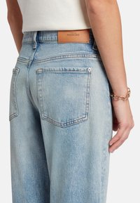Jeans de mezclilla azul claro con un corte relajado, que cuentan con bolsillos traseros y una etiqueta de marca de cuero marrón en la cintura. Detalles de costura mínimos.