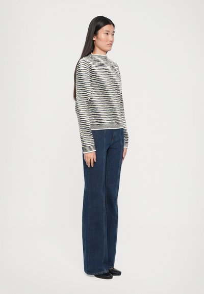 Črtasta pletena jopica v črni, kremni in bež barvi z visokim ovratnikom; kombinirana s temno modrimi širokimi jeans hlačami. Model stoji v profilu pred belo ozadjem.