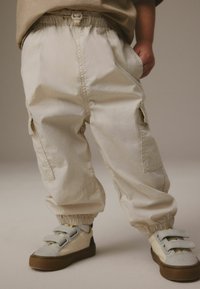 Beige cargo bukser med elastiske manchetter, der har sidelommer og justerbar talje, parret med grå og creme sneakers på en lys baggrund.