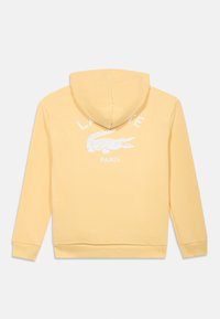 Κίτρινο φούτερ με κουκούλα από βαμβάκι. Στο μπροστινό μέρος φέρει λευκό κείμενο "LACOSTE PARIS" και γραφικό με κροκόδειλο. Μαλακή υφή, ριμπ μανσέτες και τελείωμα.