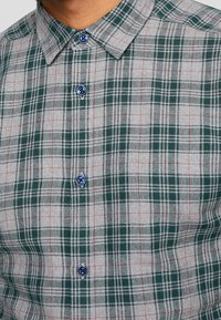 Geruite shirt met een groen, grijs en rood ruitpatroon; heeft een kraag en blauwe knopen. Gemaakt van een geweven stof met een gladde textuur.