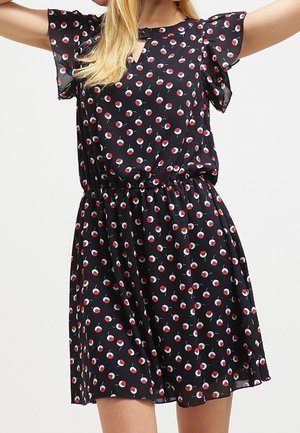 Kobieta ubrana w czarną sukienkę do kolan z falbanowymi rękawami i czerwono-białym abstrakcyjnym wzorem w kształcie kół.