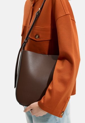 Osoba w pomarańczowej kurtce trzyma ciemnobrązową skórzaną torbę na ramię z regulowanym paskiem, ubrana również w jasnoniebieskie dżinsy.