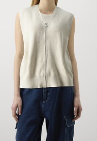 Helle beige ärmellose Strickjacke mit geripptem Muster, ausgestattet mit einem Reißverschluss vorne und einem entspannten Schnitt, kombiniert mit dunkelblauen Cargo-Hosen.