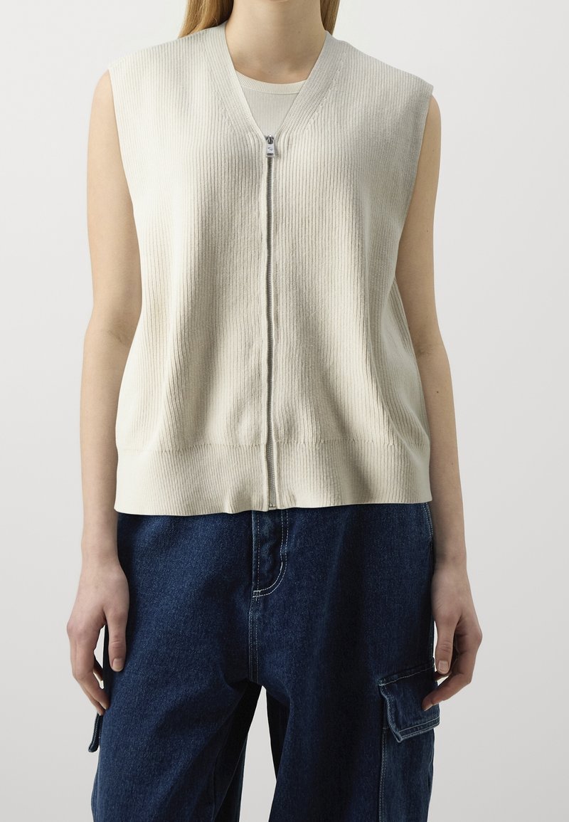 Helle beige ärmellose Strickjacke mit geripptem Muster, ausgestattet mit einem Reißverschluss vorne und einem entspannten Schnitt, kombiniert mit dunkelblauen Cargo-Hosen.