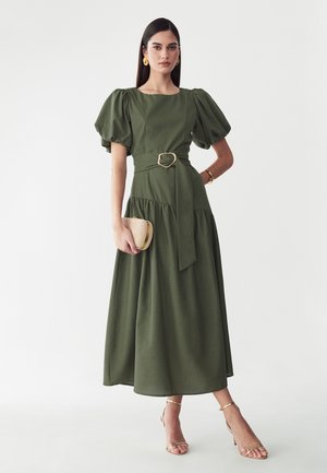 Mujer con vestido midi verde oliva de mangas abullonadas con cinturón, lleva un bolso de mano beige y sandalias y pendientes dorados.