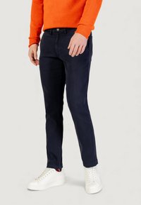Pantaloni chino in cotone blu navy con vestibilità slim, abbinati a sneaker in pelle bianca. Il modello indossa un maglione arancione a maglia con polsini a costine.