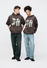 Hanorac gri închis cu glugă, imprimeu grafic cu fețe, buzunar frontal și croială lejeră. Purtat cu blugi largi din denim și pantofi negri masivi.