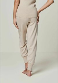Beige, gerippte Jogginghosen mit schmal zulaufendem Knöchel und elastischen Bündchen. Weicher Stoff, figurbetontes Design, barfuß getragen, von hinten betrachtet.