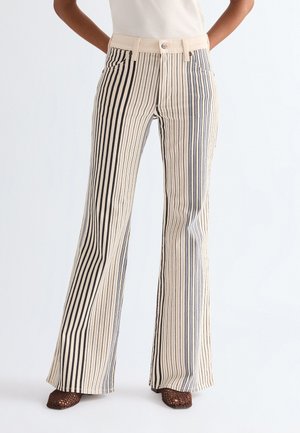 Κάτω μέρος του σώματος ατόμου που φοράει παντελόνι με ψηλή μέση και φαρδιά μπατζάκια σε κατακόρυφες μαύρες και μπεζ ρίγες, σε συνδυασμό με καφέ πλεκτά παπούτσια με ανοιχτά δάχτυλα.