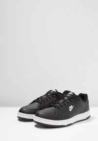 Zapatillas deportivas negras de cuero con una textura suave. Incluyen una suela blanca, cordones negros y un logotipo blanco prominente en el costado.