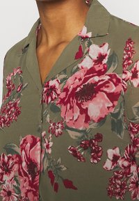 Blommig blus i olivgrön tyg med stora mönster av rosa och röda blommor, med krage och knappdetaljer fram.