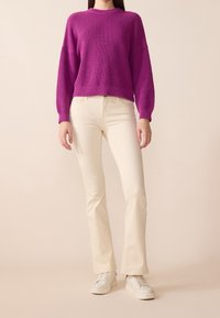 Fioletowy sweter dzianinowy o prążkowanej strukturze, w połączeniu z kremowymi dopasowanymi spodniami w kształcie dzwonów. Modelka nosi białe trampki.