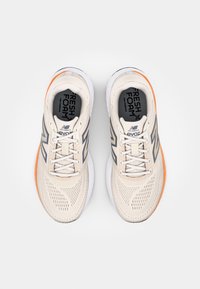 Beige løbesko med mesh-materiale, der har grå accenter, en tyk hvid mellemsål og orange detaljer ved hælen. Snørebåndsdesign.