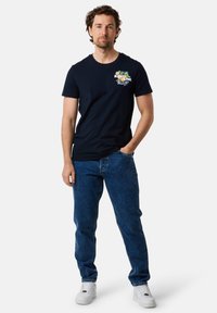 Marineblåt t-shirt med et farverigt grafisk print på brystet, parret med blå denimjeans og hvide sneakers. Casual pasform, korte ærmer.