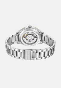 Zegarek ze stali nierdzewnej z polerowanym wykończeniem, okrągłą kopertą oraz przezroczystym tyłem, który ukazuje mechanizm. Zawiera bezpieczny zamek.