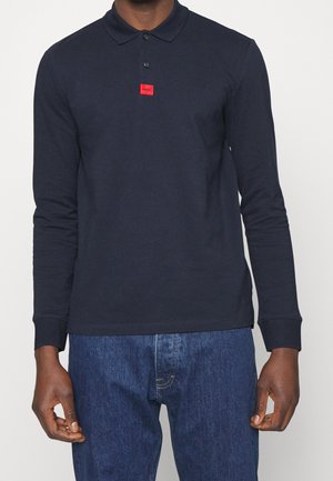 Mann trägt ein langärmeliges dunkelblaues Polohemd mit einem kleinen roten Quadrat-Logo auf der Brust und blaue Jeans, steht vor einem schlichten Hintergrund.