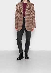 Blazer oversize marron à carreaux avec deux boutons, porté sur un pull côtelé bordeaux et accompagné d'un jean noir droit et de bottes noires.