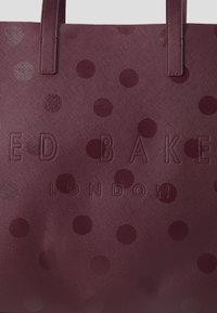 Bordeaux tote bag met een textuur, met een patroon van gegolfde stippen en het logo "RED BAKE LONDON" subtiel geïntegreerd.
