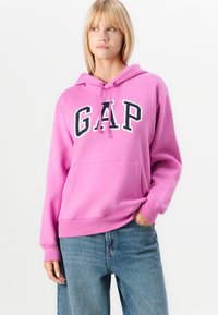 Różowa bluza z kapturem z kangurzą kieszenią, z dużym, wyraźnym logo "GAP" w białym i granatowym. Kaptur z trokami; ściągacze przy mankietach i u dołu.