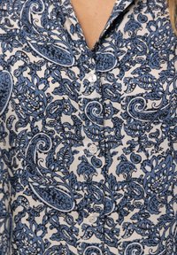 Camicia blu e bianca con bottoni, caratterizzata da un motivo paisley, tessuto testurizzato e piccoli bottoni bianchi lungo la parte anteriore.
