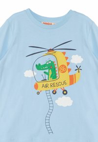 T-shirt azzurro chiaro con un pilota coccodrillo in stile cartone animato a bordo di un elicottero giallo, con la scritta "SALVATAGGIO AEREO" e grafiche di nuvole.