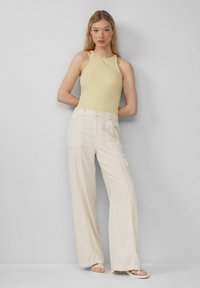Lichtgele geribde tanktop en wijdvallende crèmekleurige broek met zijzakken, gecombineerd met witte sandalen met banden, op een neutrale achtergrond.