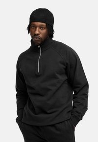 Schwarzer Sweatshirt mit halbem Reißverschluss am Kragen, gerippten Bündchen und Raglanärmel. Glatte Textur und einfaches Design. Das Modell trägt eine passende Mütze.