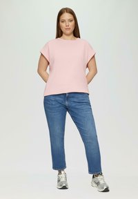 Helles rosa Kurzarmhemd mit entspannter Passform, kombiniert mit blauen, kurz geschnittenen Jeans. Die Schuhe sind metallisch silberne Sneakers. Einfaches, sauberes Design.