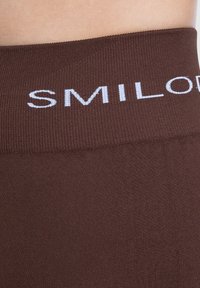 Leggings marroni con una vita lavorata a maglia con la scritta "SMILODON" in lettere bianche. La texture liscia mette in risalto un design elegante.