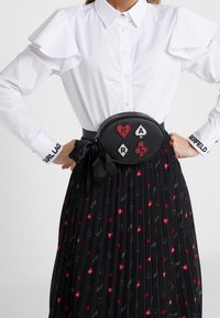 Svart läder midjeväska med hjärt- och kortsymboler i röd och silver, bärs med en vit skjorta och en plisserad svart kjol med färgglada mönster.