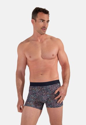 Muscular man with short curly hair modeling fitted dark boxer briefs with a colorful floral pattern, posing against a plain light background.