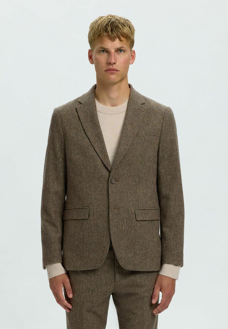 Jeune homme portant une veste de costume marron à carreaux et un pantalon assorti sur un pull beige clair, debout devant un fond clair uni.