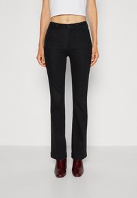 Jeans de campana negros de denim, con cremallera y cierre de botón, bolsillos frontales y una textura suave. Llevados con botas de tacón burdeos.