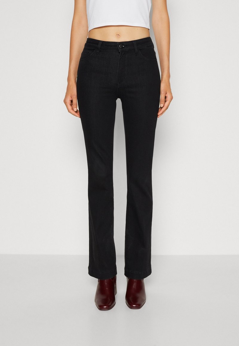 Jeans de campana negros de denim, con cremallera y cierre de botón, bolsillos frontales y una textura suave. Llevados con botas de tacón burdeos.