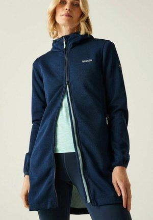 Vrouw die een lange marineblauwe hoodie met rits draagt over een lichtblauw shirt en donkere legging, staand tegen een effen achtergrond.