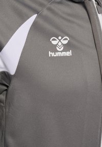 Graue Sportjacke mit weißen Akzenten, versehen mit einem weißen Hummel-Logo und einem vorderen Reißverschluss auf der rechten Seite.