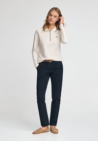 Polo shirt beige a maniche lunghe con colletto e tre bottoni, abbinato a pantaloni blu scuro e scarpe slip-on in camoscio color tan. Design semplice.