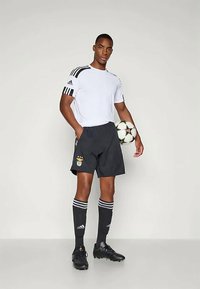 Valge lühikeste varrukatega Adidas särk mustade triipudega, mustad lühikesed püksid embleemiga, mustad sokid valgete triipudega ja mustad jalgpallijalatsid, hoides palli.