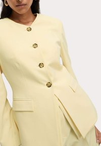 Yellow tailored blazer with a round neckline, five large decorative buttons, side pockets, and a single front flap detail.
