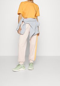 Osoba v oranžovém zkráceném tričku, smetanových teplákách s oranžovým pruhem, zelených teniskách a světle modré mikině uvázané kolem pasu.