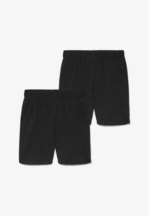 Femme debout, mains dans le dos, portant un t-shirt blanc à manches courtes, un short de cyclisme noir, des chaussettes grises et des baskets blanches sur fond blanc.