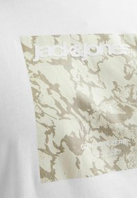 Balta marškinėliai su tekstūrizuotu, kamufliažo tipo raštu šviesiai bežine ir grietinėlės spalvomis, kuriuose ryškiai matomas "jack & jones" logotipas.