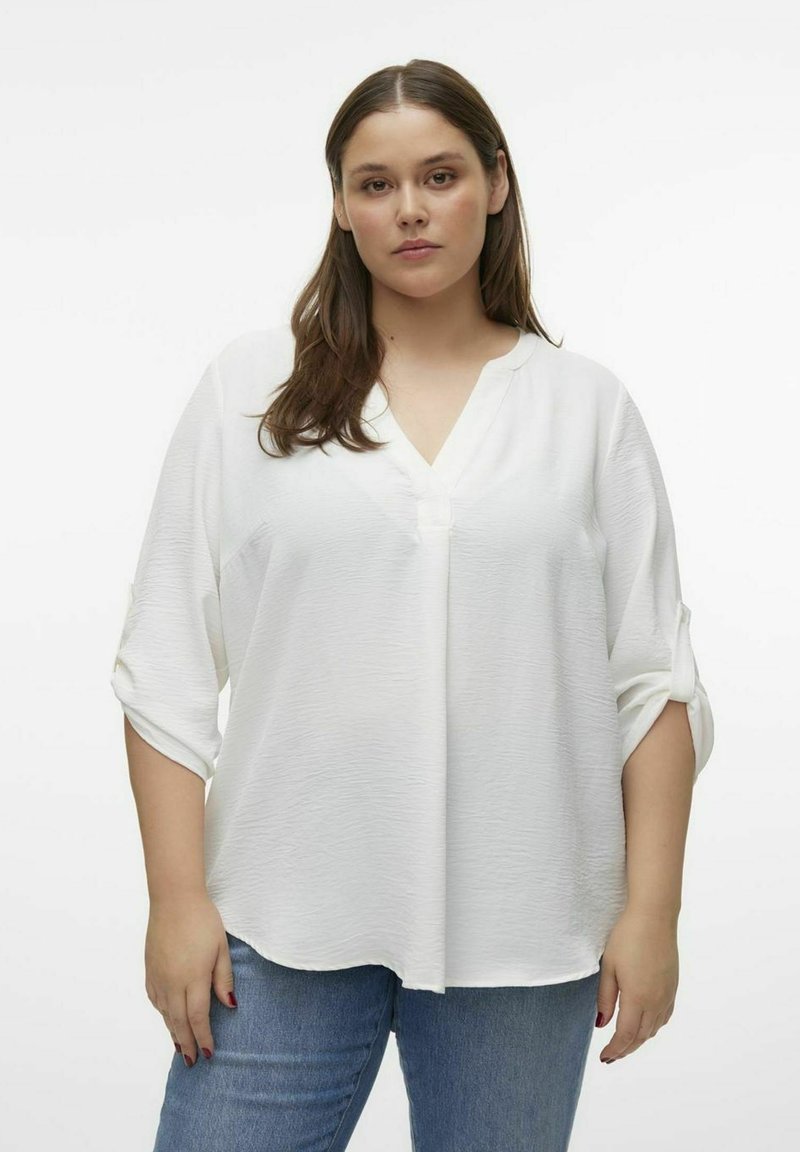 Blouse blanche avec un col en V et des manches trois-quarts. Tissu texturé, coupe ample, avec des poignets roulés et un détail de bouton au niveau du col.