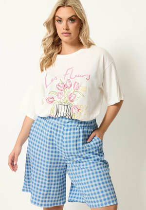 Blonde vrouw die een wit T-shirt draagt met een roze tulpafdruk en de tekst "Les Fleurs", gecombineerd met blauw-wit geruite shorts met hoge taille.