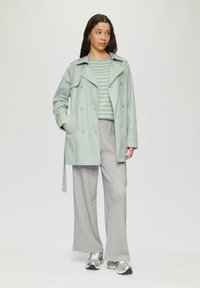 Lichtgroene trenchcoat met dubbele knopen, gedragen over een gestreepte groen-witte top en grijs geruite wijde broek. Zilveren sneakers.